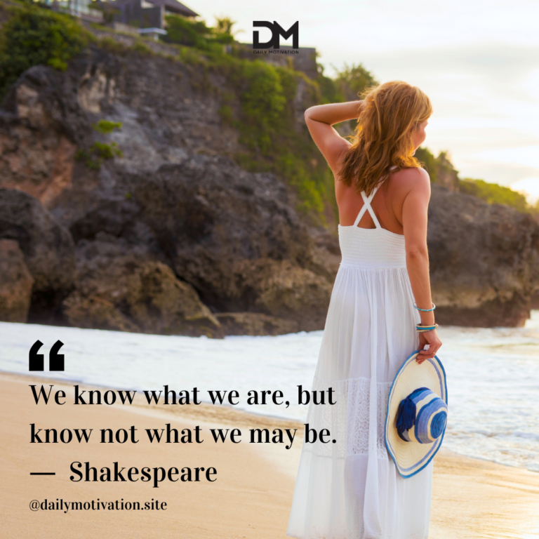 A woman in a white dress carries a hat and walks along a beach, looking out at the water.